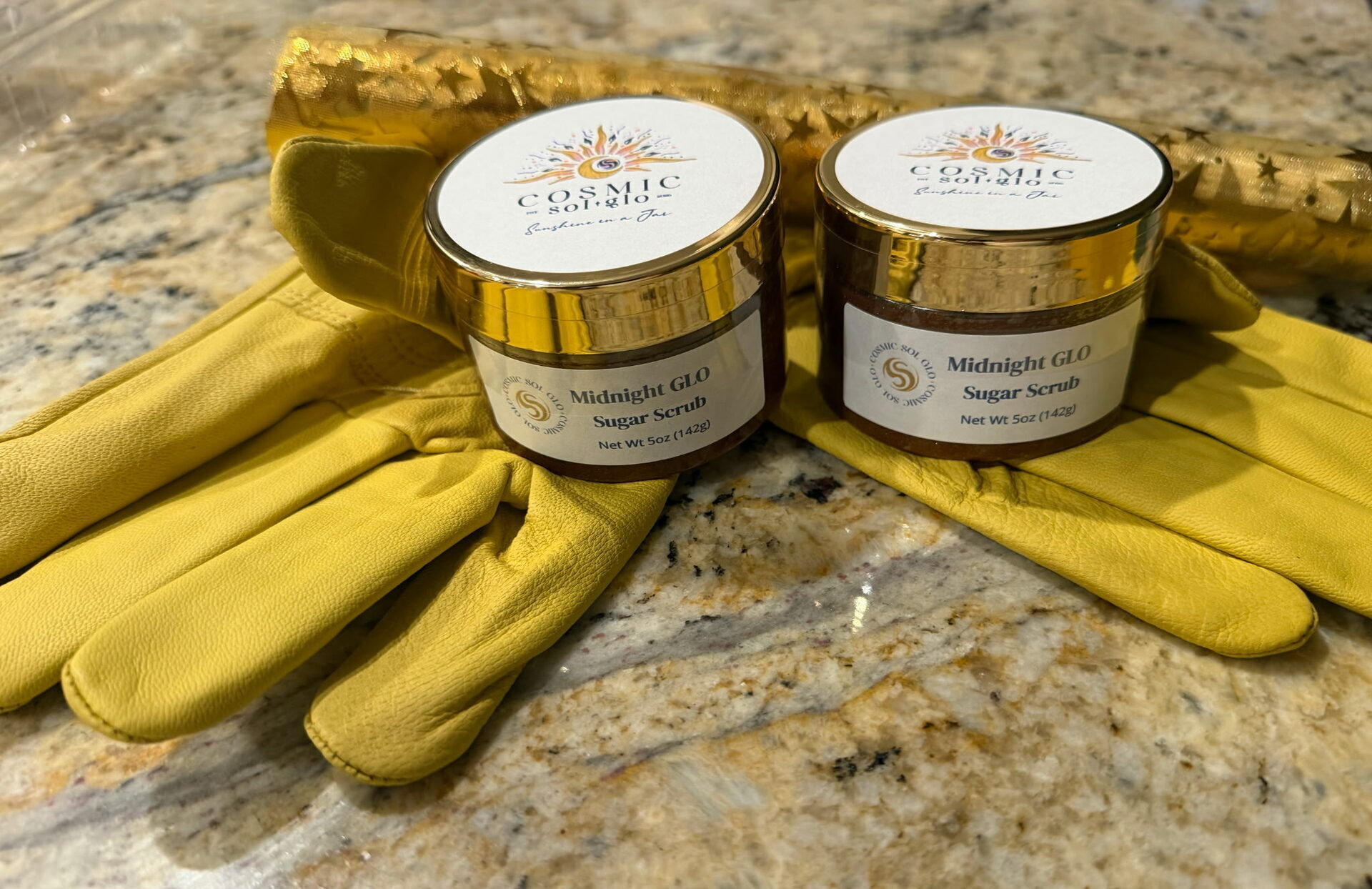 Two Midnight Glo sugar scrub jars with gold lids resting on tan work gloves, gold star gift wrap behind