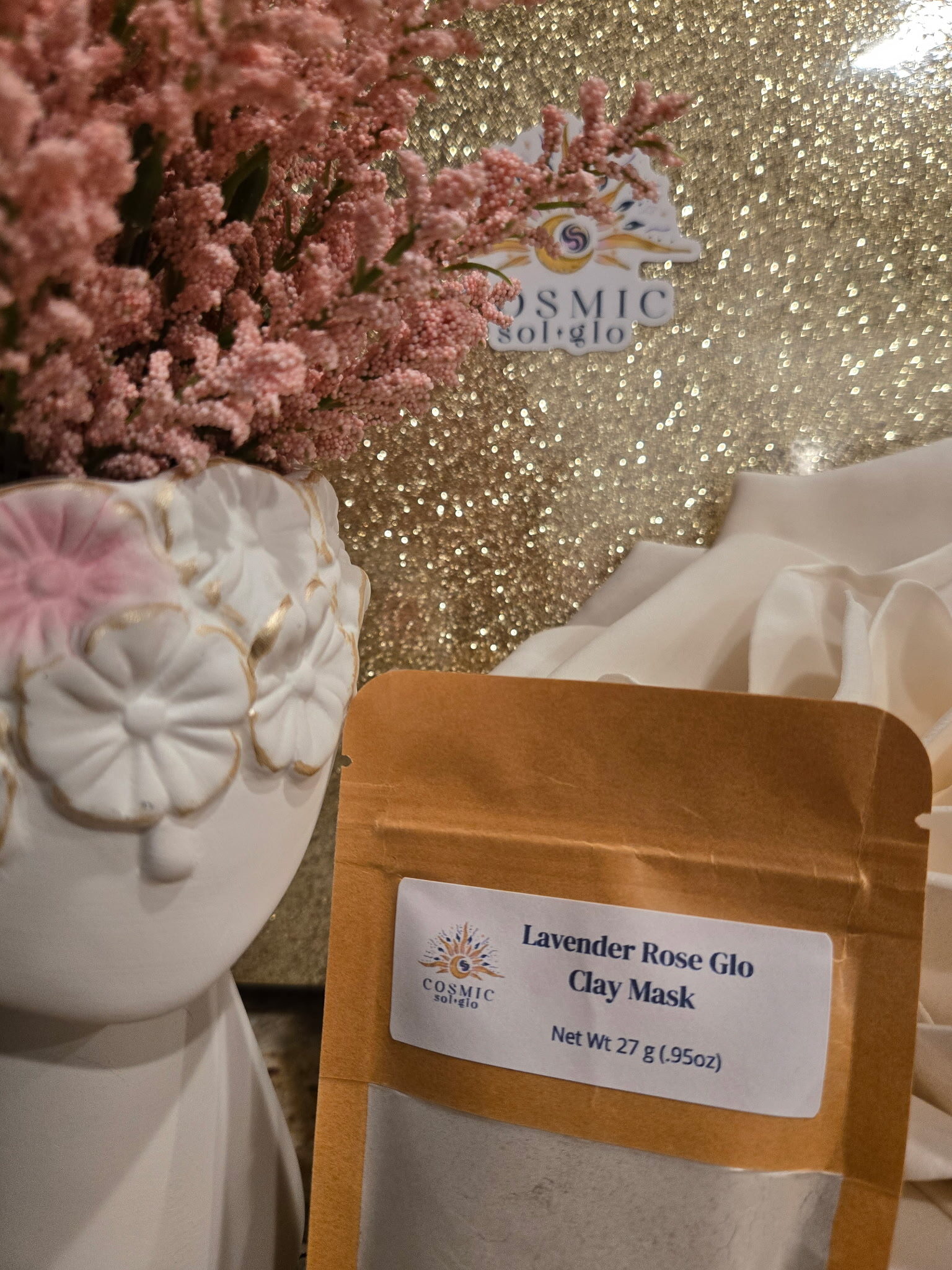Lavender Rose Glo clay mask pouch propped upright among flowers and fabric roses