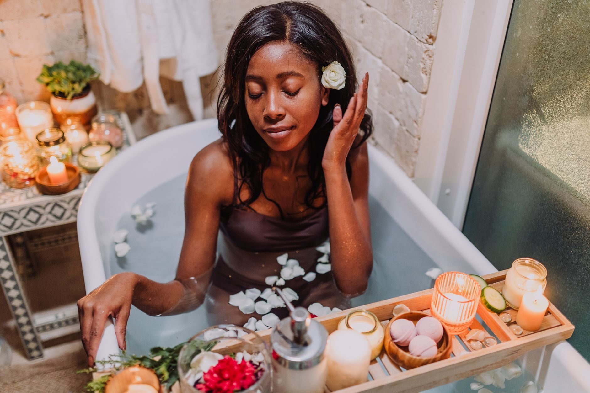 A woman resting with eyes closed in a petal-strewn bath surrounded by warm candles