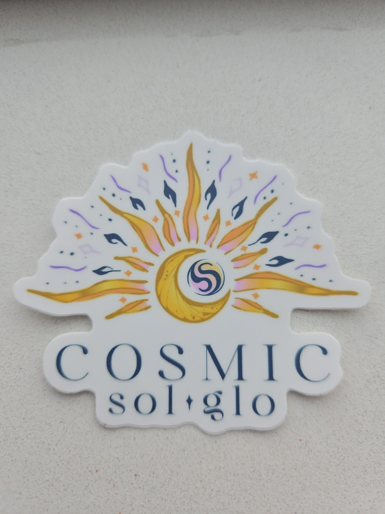 Cosmic Sol Glo sunburst logo die-cut vinyl sticker, 3 inches, mounted on a textured white wall
