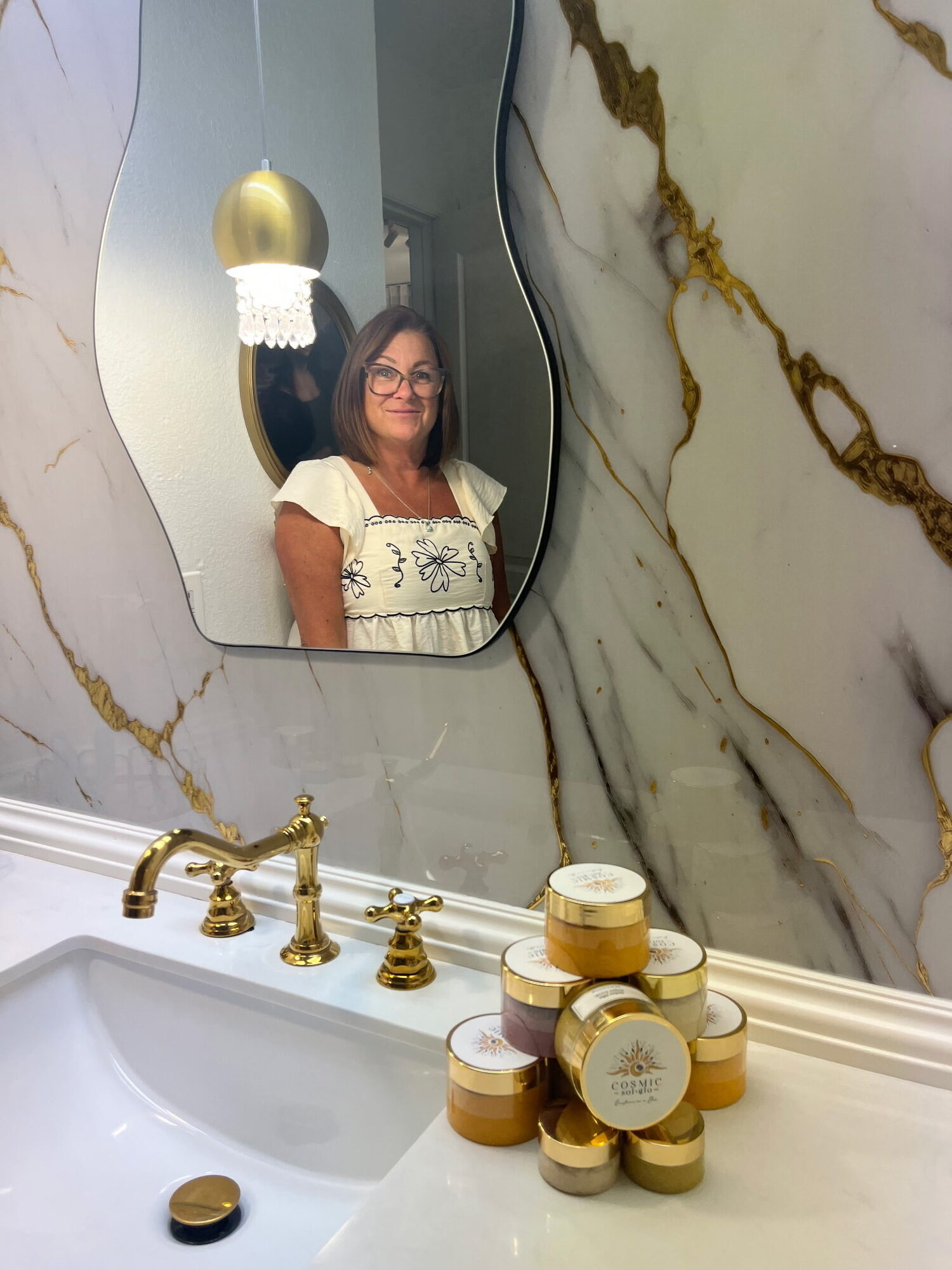 Tracy VanBuren smiling in a mirror reflection, with stacked jars of Cosmic Sol Glo scrubs on the counter in front of her