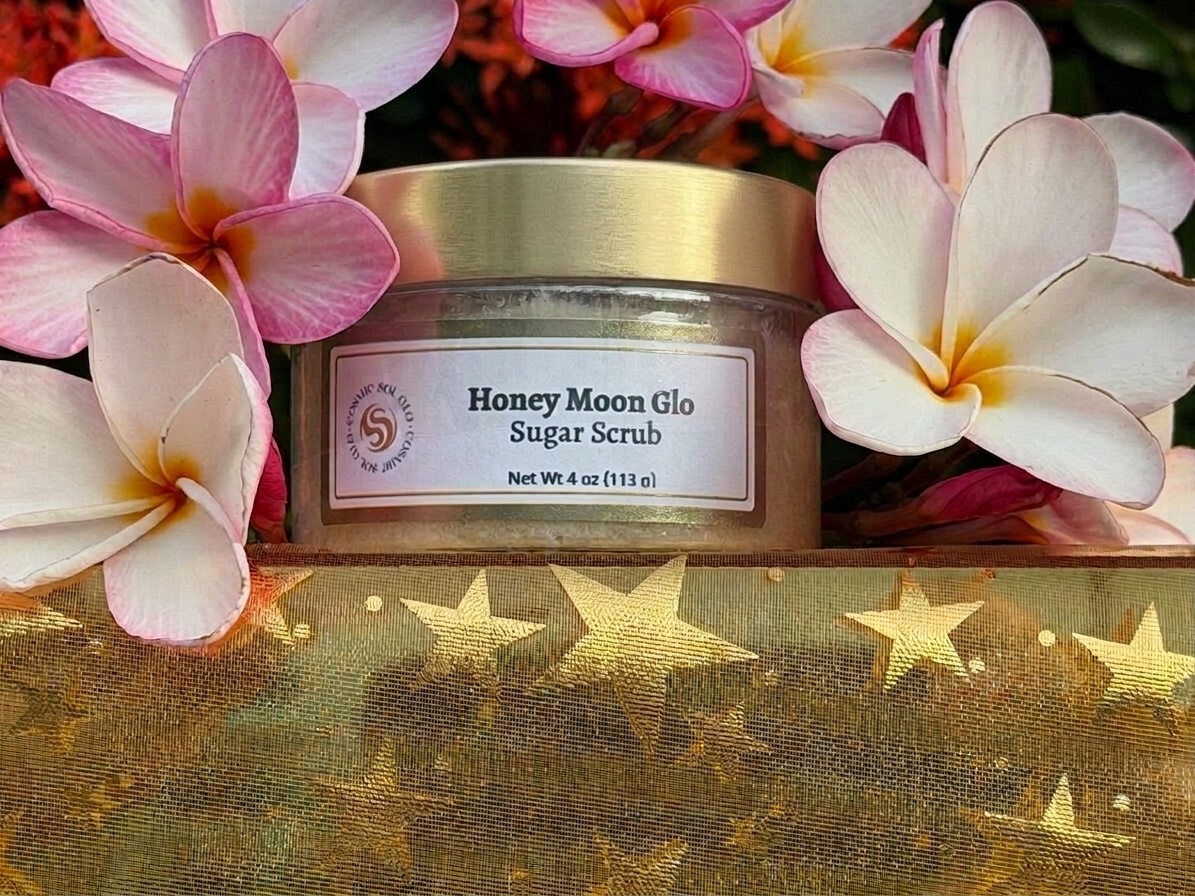 Honey Moon Glo sugar scrub jar with pale plumeria flowers over golden star fabric