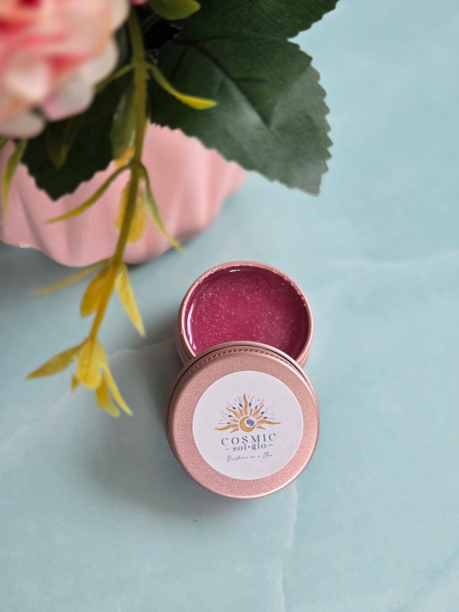 Cosmic Sol Glo Cherry Nebula lip scrub, a deep berry-toned black cherry scrub in an open rose-gold tin styled with pink florals