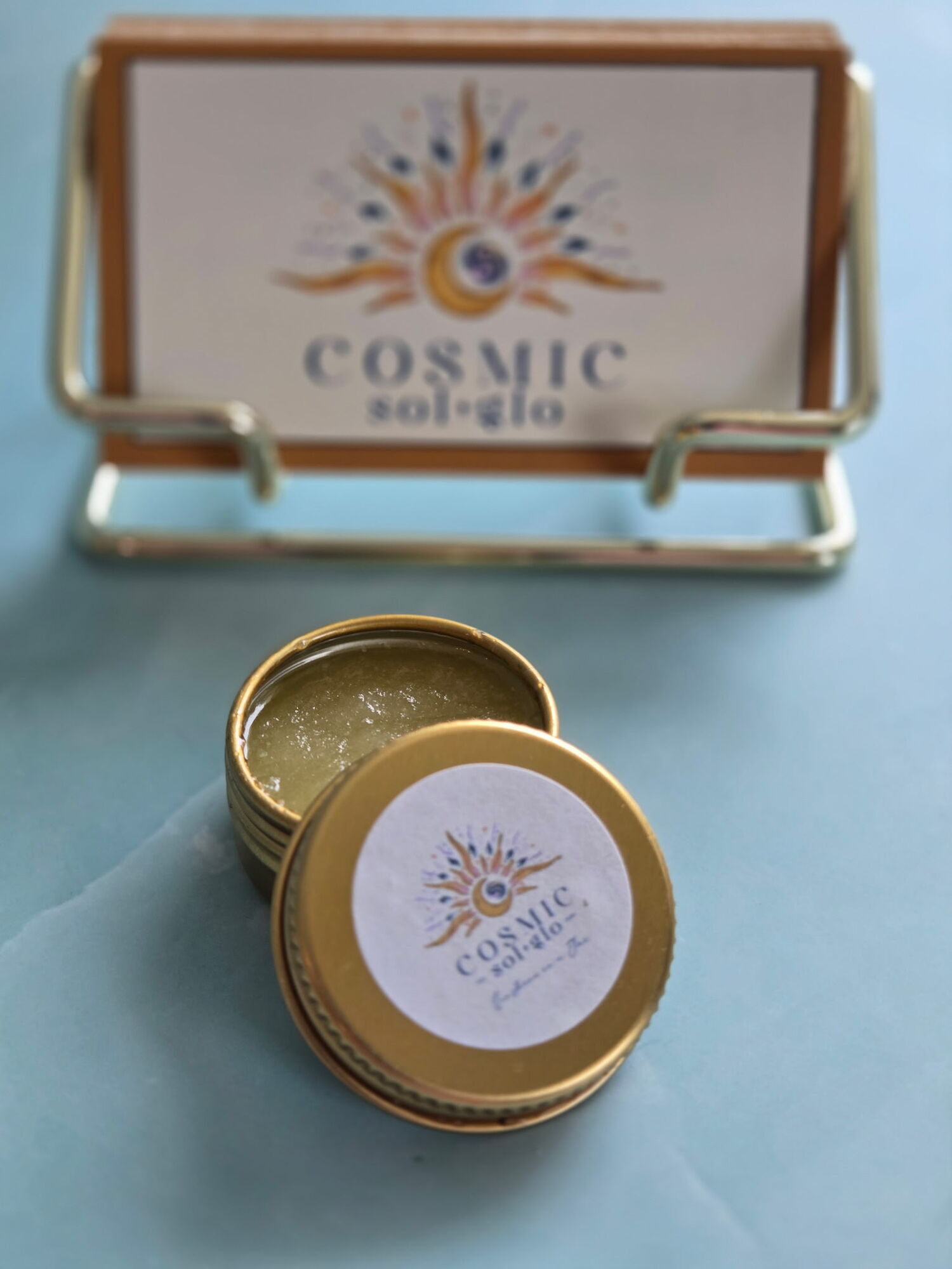 Cosmic Sol Glo Golden Aura lip scrub, a golden orange-creamsicle scrub in an open gold tin beside a Cosmic Sol Glo display card