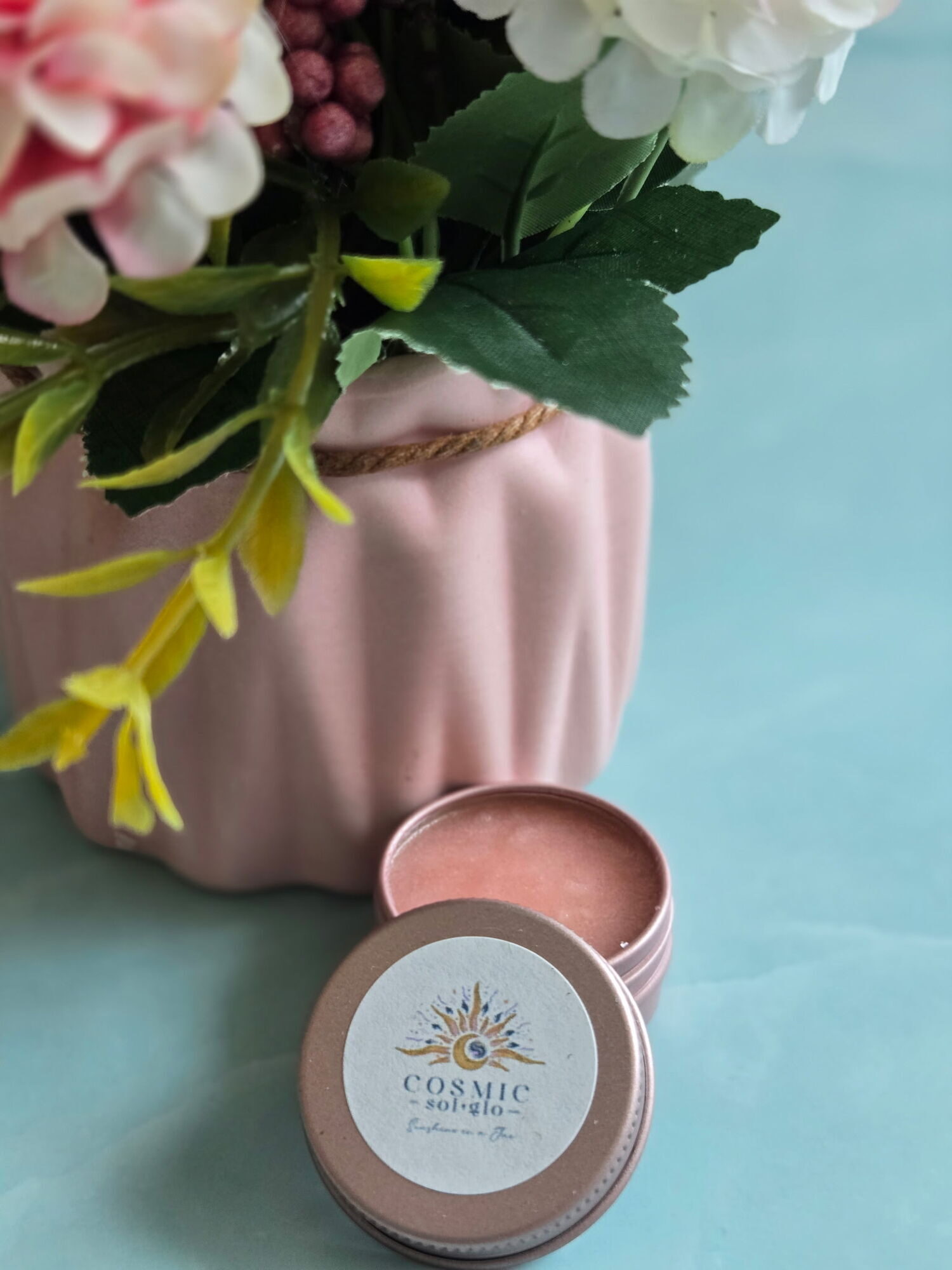 Cosmic Sol Glo Tropical Nova lip scrub, a coral fruit-punch-toned scrub in an open rose-gold tin styled with pastel florals